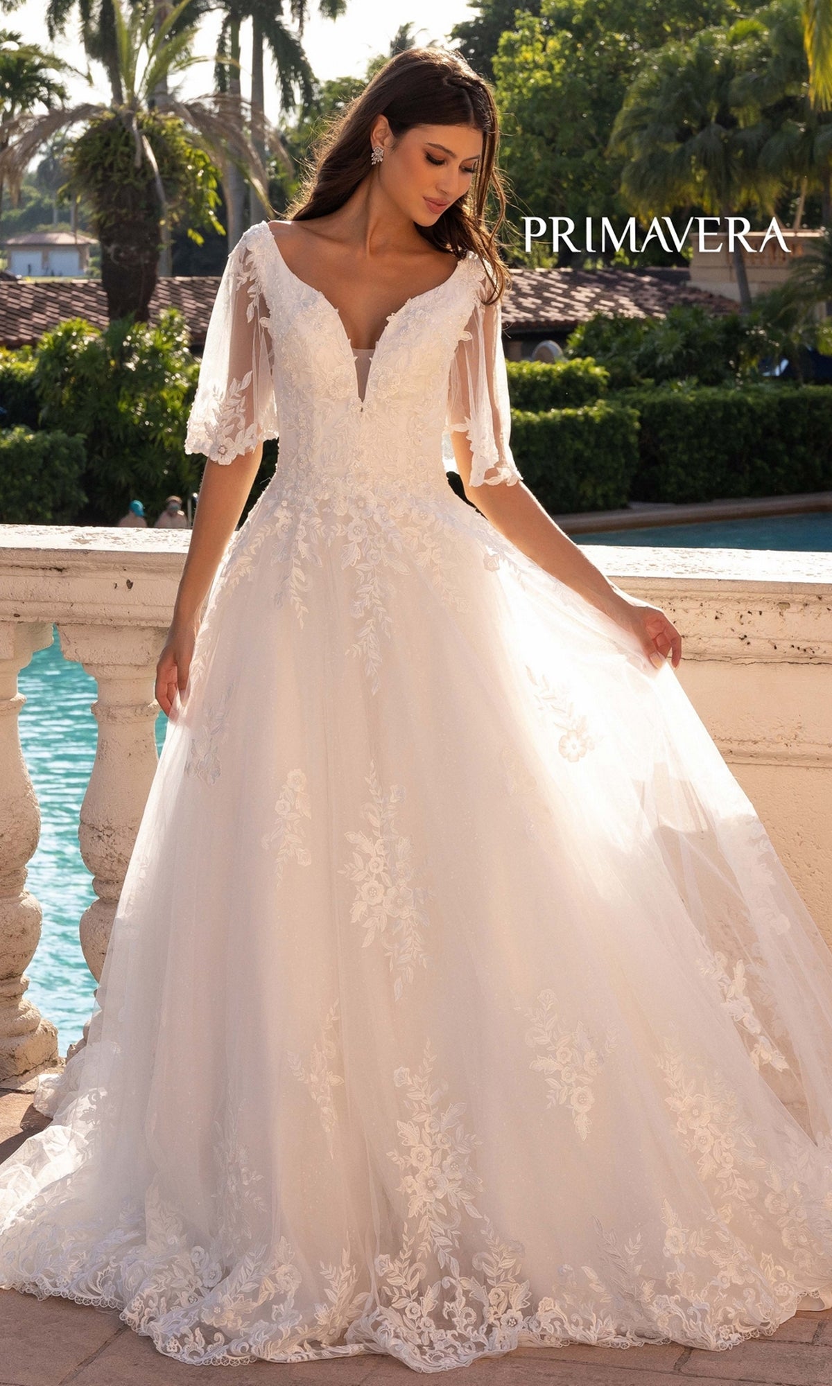 A woman stands outdoors by a stone railing, wearing the Long Wedding Dress 11134 by Primavera—an ivory A-line gown with sheer sleeves. A pool and palm trees are visible in the background.